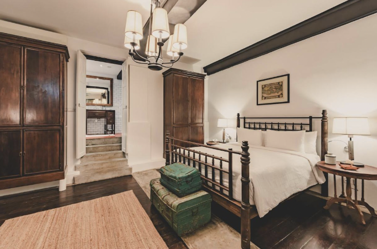 Hotel room with antique wardrobe, bed and green ottoman.